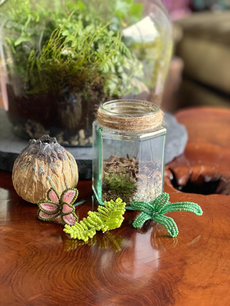 May include: A glass terrarium with greenery and a small glass jar with twine. Decorative beaded plant accents in pink, green, and a brown, textured object are arranged on a wooden surface. The image showcases a nature-inspired theme.