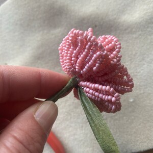 May include: A handmade pink beaded flower with a green stem and leaves. The flower is made of small, light pink beads, and the stem and leaves are a muted green colour. The flower is held in place with a wire.