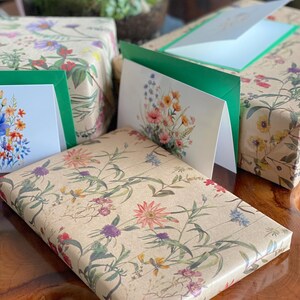 May include: Gift-wrapped packages and greeting cards with floral designs. The wrapping paper and cards feature a beige background with colorful flowers and green leaves. The cards have green borders and white interiors. The gifts are arranged on a wooden surface.