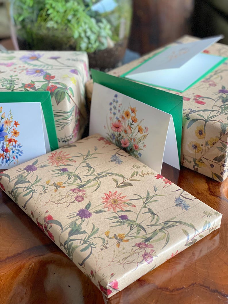 May include: Gift-wrapped packages and greeting cards with floral designs. The wrapping paper and cards feature a beige background with colorful flowers and green leaves. The cards have green borders and white interiors. The gifts are arranged on a wooden surface.