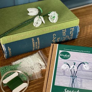 May include: A beaded snowdrop flower craft kit with a green and white color scheme. The kit includes beads, wire, and thread, along with a book titled "The Illustrated Guide to Garden Flowers". The kit is designed to create beaded snowdrop flowers.