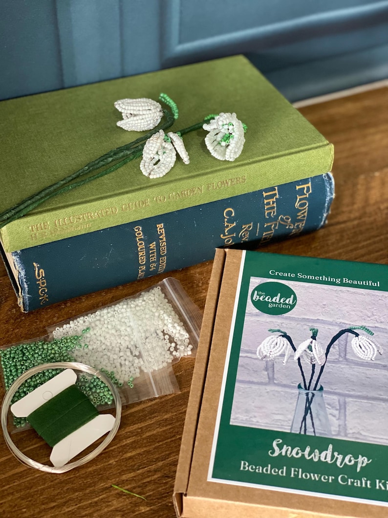 May include: A beaded snowdrop flower craft kit with a green and white color scheme. The kit includes beads, wire, and thread, along with a book titled "The Illustrated Guide to Garden Flowers". The kit is designed to create beaded snowdrop flowers.