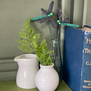 May include: A decorative display featuring two beaded dragonfly ornaments with teal and purple wings, and a green stem. The dragonflies are placed in a clear glass bottle. A small white jug and vase hold greenery. A stack of books is in the background.