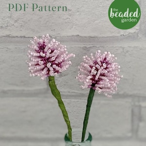 May include: Two pink and purple beaded flower stems in a clear glass vase. The flowers are in a cluster of small beads and are arranged in a vase against a white brick wall. The text "PDF Pattern" and "the beaded garden" are visible in the image.