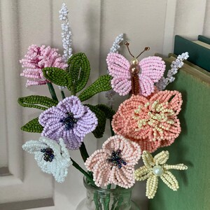 May include: A handmade beaded flower arrangement in a clear glass vase. The bouquet features pink, purple, white, and peach flowers, plus a beaded butterfly. Green leaves and stems complete the floral display.