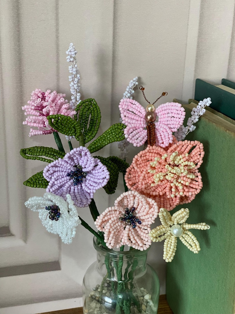May include: A handmade beaded flower arrangement in a clear glass vase. The bouquet features pink, purple, white, and peach flowers, plus a beaded butterfly. Green leaves and stems complete the floral display.