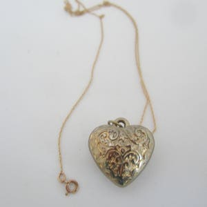 May include: A gold chain necklace with a detailed heart pendant. The heart is silver and has an intricate floral design.