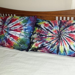 Puede incluir: Dos fundas de almohada de teñido anudado con un patrón de remolino en tonos de azul, rojo, amarillo y negro. Las fundas de almohada están sobre una cama blanca con una cabecera de madera.