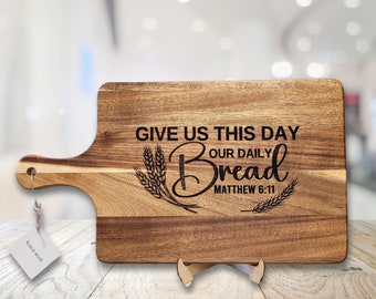 Puede incluir: Tabla de cortar de madera de acacia con asa. La tabla presenta el texto grabado "GIVE US THIS DAY OUR DAILY BREAD MATTHEW 6:11" con ilustraciones de trigo y hojas. Una pequeña etiqueta dice "Acacia wood".