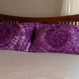 Puede incluir: Dos fundas de almohada de color morado con estampado tie-dye en una cama blanca con una cabecera de madera.