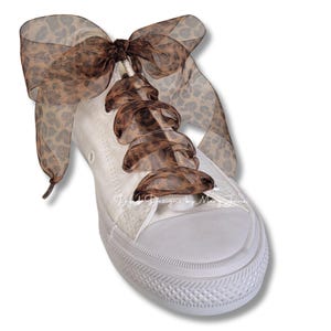 May include: White canvas sneakers with brown and tan leopard print ribbon laces and a matching bow on top.