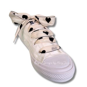 May include: White canvas sneaker with black heart-patterned ribbon laces. The shoe has a white rubber toe cap and sole. The laces are tied in a bow. The text "Designs by Mary Jane" is visible on the shoe.