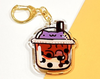 May include: A clear acrylic keychain featuring a cartoon cat sitting inside a boba tea cup. The boba tea is purple, red, and white with black boba pearls. The keychain has a gold metal ring.