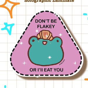 May include: A holographic sticker with a pink triangle shape and a cartoon frog wearing a brown pastry on its head. The sticker says "Don't be Flakey or I'll eat you".