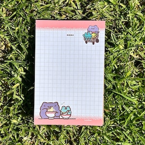 May include: A pink memo pad with a grid pattern and a cartoon illustration of a purple cat with a bowl of noodles and a green frog with a bowl of noodles. The text "memo" is printed at the top of the pad.