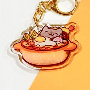 May include: A clear acrylic keychain featuring a cartoon cat in a bowl of ramen. The cat is gray with a pink nose and is surrounded by noodles, an egg, and other toppings. The keychain has a gold metal ring.