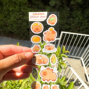 May include: A clear sticker sheet with a textured finish featuring illustrations of capybaras in various poses. The sheet includes capybaras eating corn, swimming, and relaxing. The text "Capybara Sticker Sheet - textured -" is printed on the sheet.