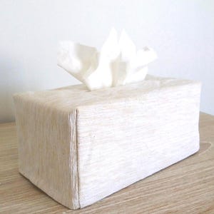 May include: A rectangular tissue box cover featuring a textured, off-white fabric finish with subtle vertical ribbing. The cover is designed to fit over a standard tissue box, with a single white tissue protruding from the top opening.