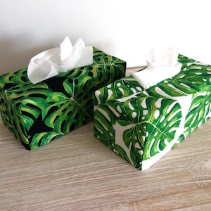 May include: Two rectangular tissue box covers featuring a vibrant green monstera leaf pattern. One cover has a black background, while the other has a white background. Both covers are shown with white tissues protruding from the top opening.