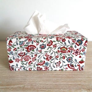 May include: A rectangular tissue box cover featuring a white background with a dense, repeating floral pattern in shades of red, blue, and green. The fabric cover is fitted over a box, with a single white tissue protruding from the top opening.
