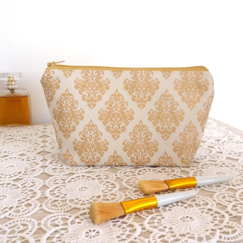 Puede incluir: Un neceser crema con un estampado damasco dorado y una cremallera dorada. Dos brochas de maquillaje con mangos blancos y detalles dorados est&aacute;n frente a la bolsa. La bolsa est&aacute; sobre una superficie de encaje blanco.