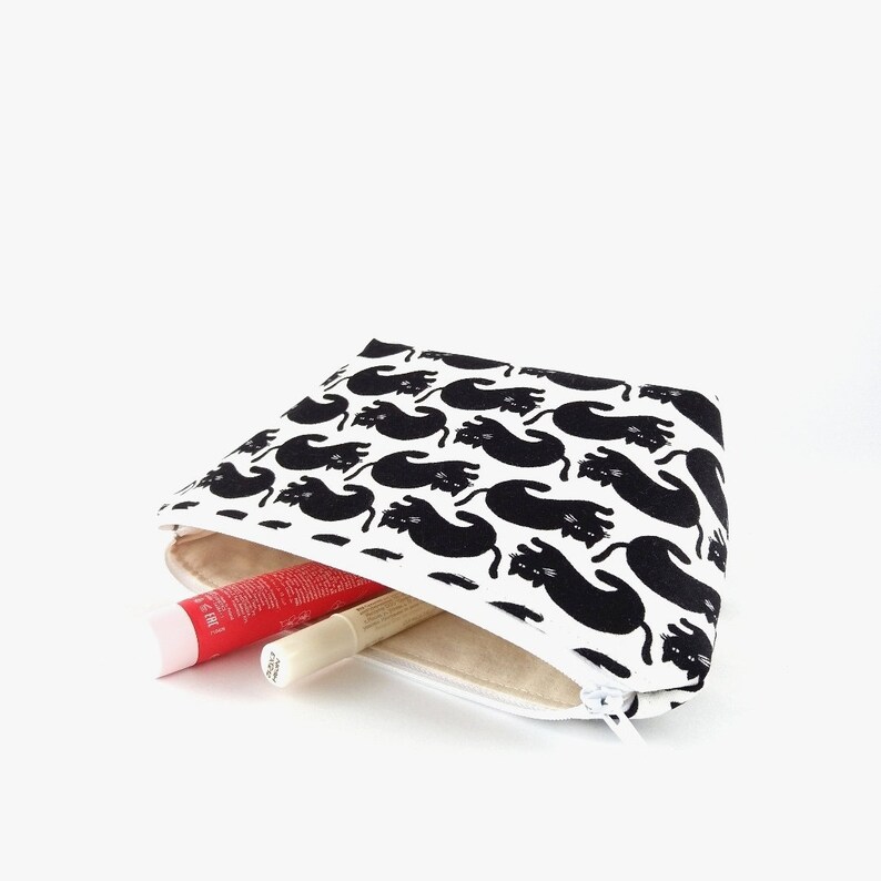 Puede incluir: Un neceser blanco con un estampado de gatos negros. La bolsa est&aacute; abierta, revelando un tubo de b&aacute;lsamo labial rojo y un tubo de brillo labial blanco. La bolsa tiene una cremallera blanca.