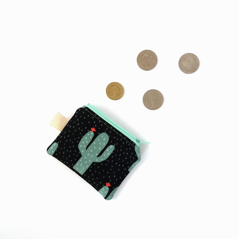 Puede incluir: Un peque&ntilde;o monedero de tela negra con un estampado de cactus en verde claro y rojo. El monedero tiene una cremallera verde claro y una etiqueta de tela beige. Varias monedas est&aacute;n esparcidas sobre un fondo blanco.