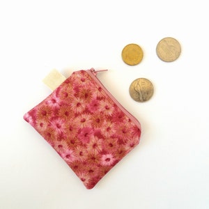 Puede incluir: Un peque&ntilde;o monedero de tela rosa con un estampado floral dorado y una cremallera rosa a juego. El monedero tiene una leng&uuml;eta de tela color crema y est&aacute; junto a tres monedas.