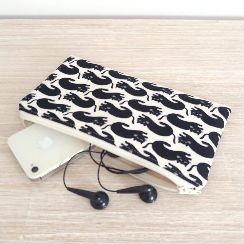 Puede incluir: Una funda rectangular con un estampado de gatos negros sobre fondo blanco, con cremallera. Se ve un smartphone blanco y unos auriculares negros. La funda est&aacute; abierta. Fondo de superficie de madera clara.
