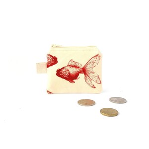 Puede incluir: Un peque&ntilde;o monedero de color crema con un estampado de pez dorado rojo. El monedero tiene un cierre de cremallera y un peque&ntilde;o lazo de tela. Varias monedas est&aacute;n esparcidas frente al monedero.