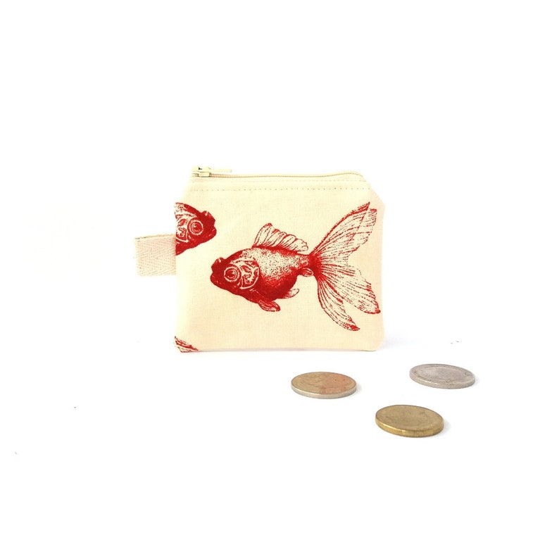 Puede incluir: Un peque&ntilde;o monedero de color crema con un estampado de pez dorado rojo. El monedero tiene un cierre de cremallera y un peque&ntilde;o lazo de tela. Varias monedas est&aacute;n esparcidas frente al monedero.