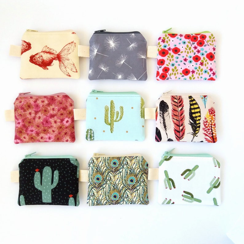 Puede incluir: Nueve peque&ntilde;os monederos cuadrados con varios dise&ntilde;os. Los dise&ntilde;os incluyen un pez dorado, dientes de le&oacute;n, flores rojas, flores rosas, un cactus, plumas, un cactus negro, plumas de pavo real y cactus verdes. Cada monedero tiene una cremallera y un peque&ntilde;o lazo de tela.