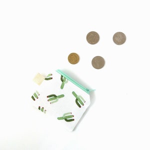 Puede incluir: Un peque&ntilde;o monedero blanco con cremallera con un estampado de cactus verde y una cremallera verde claro. Cuatro monedas est&aacute;n dispersas alrededor del monedero sobre un fondo blanco. El monedero tiene una peque&ntilde;a etiqueta de tela beige.