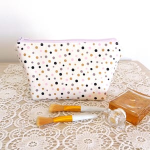 Puede incluir: Un neceser blanco con un estampado de lunares en negro, dorado, gris y rosa. La bolsa tiene una cremallera lavanda y est&aacute; junto a dos brochas de maquillaje y un frasco de perfume.