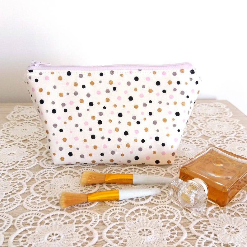 Puede incluir: Un neceser blanco con un estampado de lunares en negro, dorado, gris y rosa. La bolsa tiene una cremallera lavanda y est&aacute; junto a dos brochas de maquillaje y un frasco de perfume.