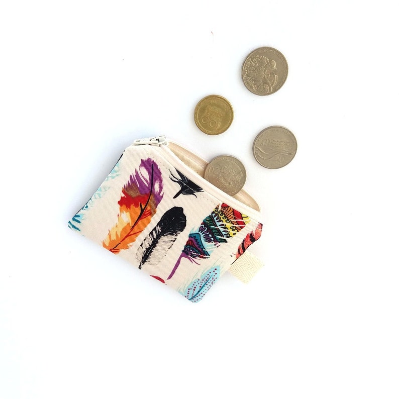 Puede incluir: Un peque&ntilde;o monedero crema con cierre de cremallera. El monedero est&aacute; decorado con coloridos dise&ntilde;os de plumas en tonos morado, naranja, negro y azul. Varias monedas est&aacute;n esparcidas alrededor del monedero sobre un fondo blanco.