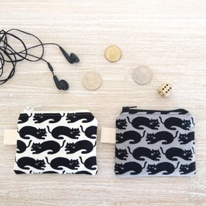 Puede incluir: Dos peque&ntilde;os monederos con cremallera y estampados de gatos. Uno es blanco con gatos negros, el otro es gris con gatos negros. Auriculares negros, monedas y un dado tambi&eacute;n est&aacute;n sobre una superficie de madera clara.