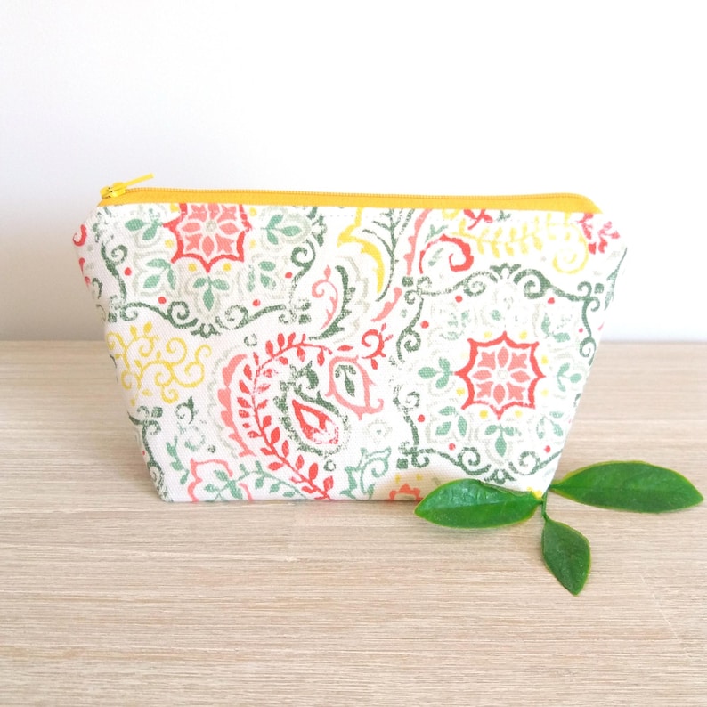 Puede incluir: Un peque&ntilde;o neceser rectangular con cremallera amarilla. La bolsa presenta un fondo blanco con un colorido estampado de cachemira en rojo, verde, amarillo y rosa. Est&aacute; sobre una superficie de madera.