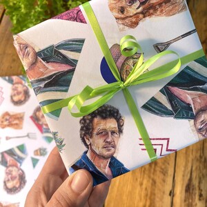 May include: Gift-wrapped present with a white background featuring illustrations of a man, a dog, and other objects. The gift is tied with a bright green ribbon. The wrapping paper has a collage-like design with various images.