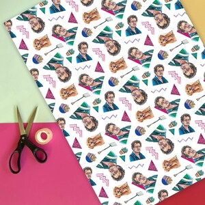 May include: White wrapping paper with a repeating pattern of a man's portrait, a dog, a gardening fork, a potted succulent, and geometric shapes in pink, green, and blue. A pair of black scissors and tape are on a pink and yellow background.