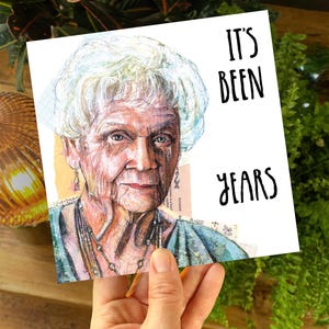 May include: A square greeting card featuring a watercolour portrait of an elderly woman. The card has the text "IT'S BEEN YEARS" in black. The woman has white hair and is wearing a blue top.