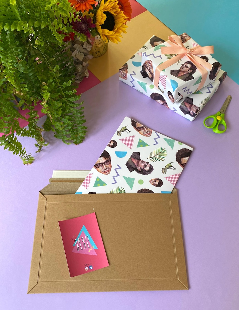 May include: Two gift-wrapped presents with a peach ribbon, patterned with faces, geometric shapes, and dinosaurs. A brown envelope contains a card with "MAIL DEAL" and a matching patterned sheet. Green scissors and flowers are also visible.