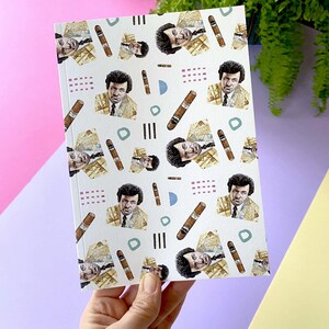May include: A white notebook featuring a repeating pattern of a man with an afro wearing a yellow jacket and holding a cigar. The background is white with geometric shapes in pink, blue, and green.