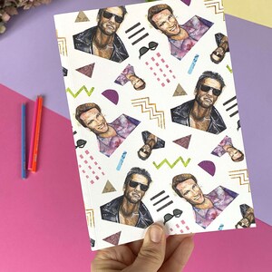 May include: A white notebook with a repeating pattern of a man's face wearing sunglasses and a leather jacket. The background is white with geometric shapes in pink, purple, green, and gold.