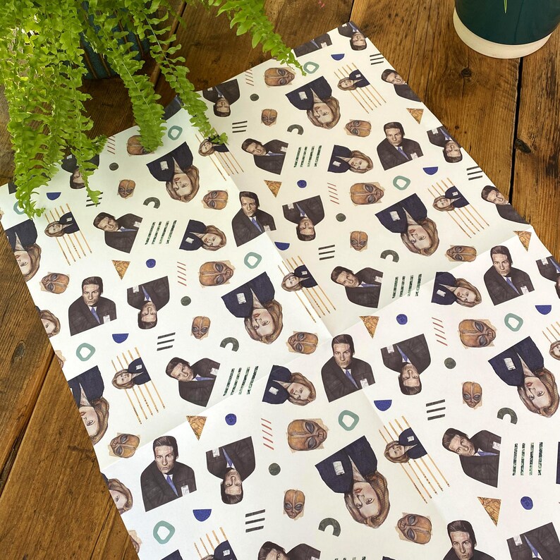 May include: White wrapping paper with a repeating pattern of a man and woman's faces, geometric shapes, and alien-like figures. The faces are of a man and woman with dark hair and dark clothing.