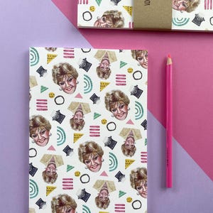 May include: A white notebook with a repeating pattern of a woman's face, a typewriter, and geometric shapes in pink, yellow, green, and black. The notebook cover has the text "DESIGNED IN 1983, 2017" and "RULED AS NOTEBOOK".
