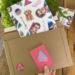 May include: A brown kraft paper envelope with a pink card and sticker featuring the text "ANGIE BEAL" in a retro design. Inside, a patterned card with illustrations of a dog, a man, and geometric shapes. A wrapped gift is also visible.