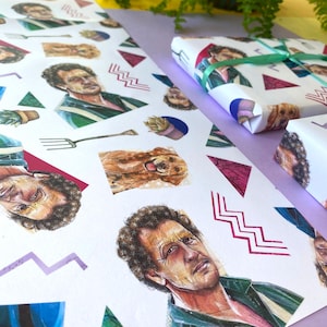 Könnte beinhalten: Weißes Geschenkpapier mit einem sich wiederholenden Muster aus einem Männerporträt, einem Golden Retriever und geometrischen Formen. Ein verpacktes Geschenk ist mit einem grünen Band gebunden. Das Papier liegt auf einer violetten Oberfläche.