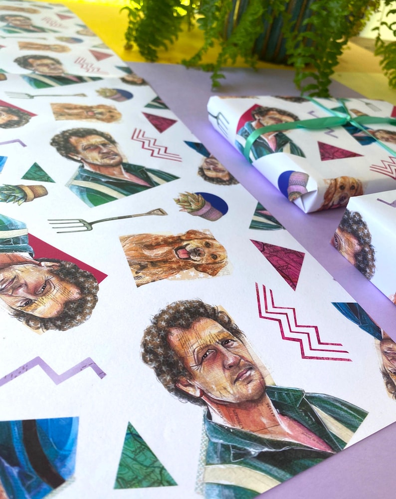 May include: White wrapping paper featuring a repeating pattern of a man's portrait, a golden retriever, and geometric shapes. A wrapped gift is tied with a green ribbon. The paper is on a purple surface.