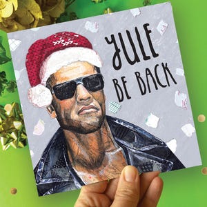 May include: Greeting card with a portrait of a man wearing a Santa hat and sunglasses. The text "Yule Be Back" is written in black script on a gray background. The card is held against a green background with gold confetti.